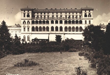 Ein historisches Hotelgebäude mit mehreren Balkonen, umgeben von einem gepflegten Garten. Die Architektur ist prächtig, mit großen Fenstern und einem eleganten Erscheinungsbild. Der Himmel ist klar, was eine einladende Atmosphäre schafft.