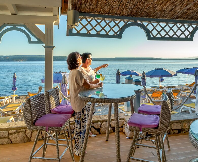 Zwei Frauen stehen an einem Tisch mit Blick auf das Meer, umgeben von Liegen und Sonnenschirmen. Die Atmosphäre ist entspannt und einladend, mit sanften Wellen im Hintergrund und warmem Licht der Abendsonne.