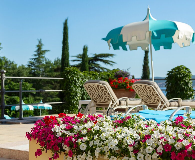 Ein sonniger Poolbereich mit Liegen und einem bunten Sonnenschirm. Bunte Blumen in roten und weißen Tönen umrahmen die Terrasse, während im Hintergrund grüne Bäume und ein Blick auf das Wasser zu sehen sind.