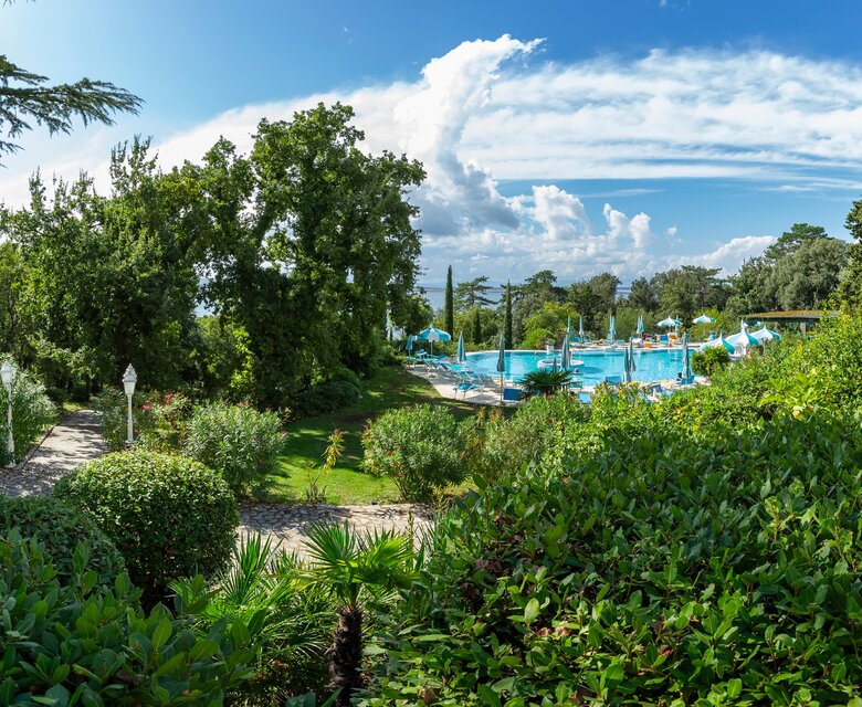 Blick auf einen gepflegten Garten mit üppigem Grün und Sträuchern, im Hintergrund ein einladender Poolbereich mit Sonnenschirmen und Liegen, unter einem strahlend blauen Himmel mit einigen Wolken.