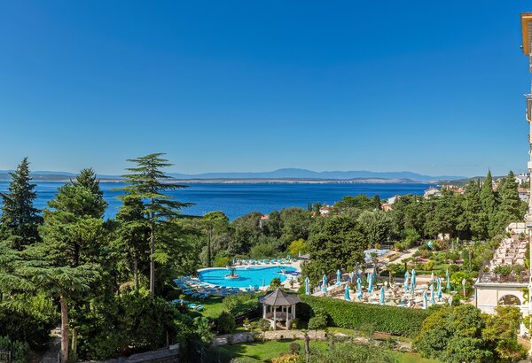 Blick auf einen Hotelgarten mit einem großen Pool, umgeben von grünen Bäumen und Liegen. Im Hintergrund erstreckt sich der See mit Bergen am Horizont unter einem klaren blauen Himmel. Doppelzimmer im Hotel direkt am Meer, Crikvenica