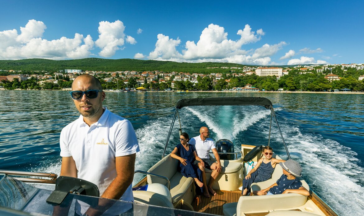 Eine Gruppe von fünf Personen fährt auf einem Boot über das Wasser. Der Kapitän steuert das Boot, während die anderen Passagiere entspannt sitzen und die Aussicht genießen. Im Hintergrund sind grüne Hügel und ein klarer blauer Himmel mit einigen Wolken zu sehen.