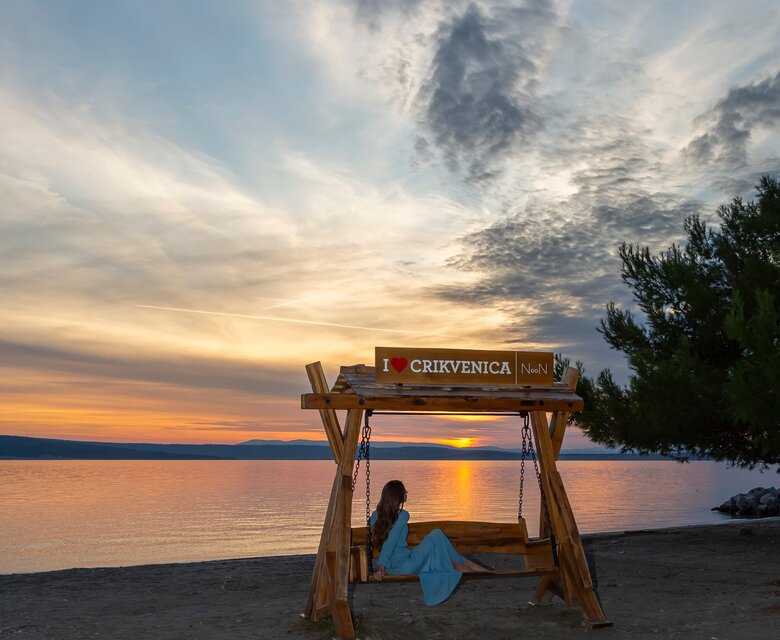 Eine Person sitzt auf einer Holzschaukel am Strand von Crikvenica, während die Sonne untergeht. Der Himmel ist mit Wolken in sanften Farben gefüllt, und das ruhige Wasser reflektiert das Licht des Sonnenuntergangs.