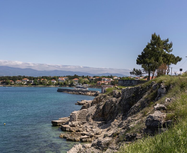 Küstenansicht mit klarem Wasser, Felsen und einer grünen Wiese. Im Hintergrund sind Häuser und ein kleiner Hafen zu sehen, unter einem strahlend blauen Himmel.