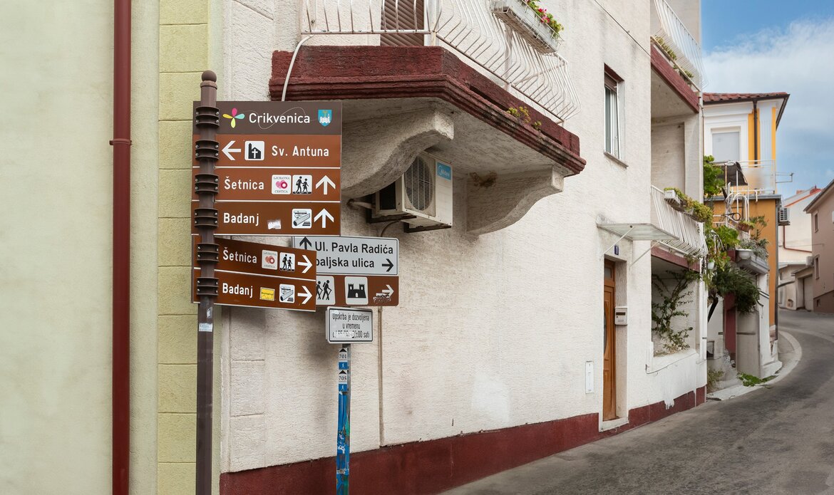 Ein Straßenschild an einer Ecke zeigt verschiedene Richtungen und Ziele in der Stadt, darunter Crikvenica und Badanj. Im Hintergrund ist ein Wohngebäude mit einem Balkon zu sehen. Die Straße verläuft sanft um die Ecke.