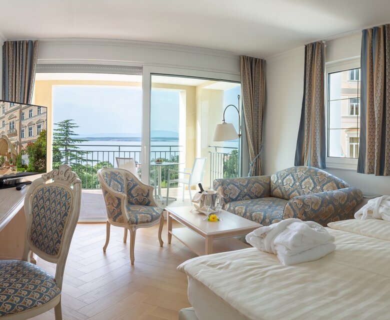 Helles Hotelzimmer mit zwei Betten, einem Sofa und einem Tisch. Ein Fernseher steht auf einem Schreibtisch. Große Fenster bieten einen Blick auf das Meer und einen Balkon. Die Einrichtung ist modern und einladend.