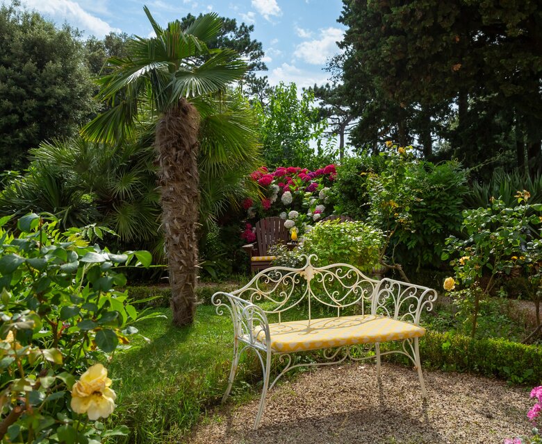 Ein einladender Garten mit üppigem Grün, bunten Blumen und einem weißen, geschwungenen Gartenbank. Palmen und Sträucher umrahmen die Szene, während die Sonne durch die Wolken scheint und eine entspannte Atmosphäre schafft.