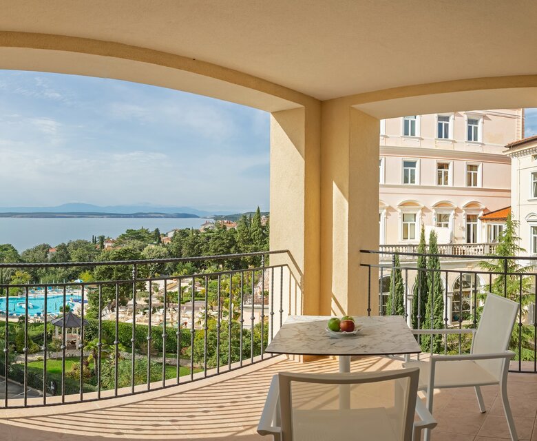 Blick von einem Balkon auf einen Pool und eine grüne Gartenlandschaft, mit dem Meer im Hintergrund. Ein Tisch mit zwei Stühlen steht auf dem Balkon, dekoriert mit einer Schale Obst. Die Atmosphäre ist entspannt und einladend.