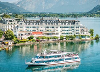Ein elegantes Hotel am Ufer eines klaren Sees, umgeben von Bergen. Ein Ausflugsboot mit Passagieren fährt gemächlich über das Wasser. Die Szenerie strahlt Ruhe und natürliche Schönheit aus.