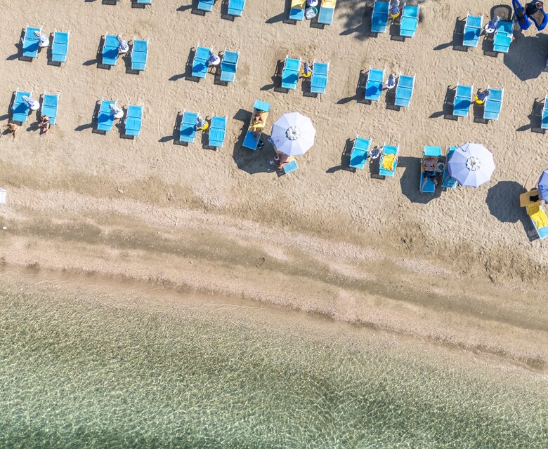 Ein Blick von oben auf einen Strand mit vielen blauen Liegen und Sonnenschirmen. Das Wasser ist klar und sanft, während einige Gäste am Strand entspannen. Im Hintergrund sind Bäume und eine Terrasse zu sehen.