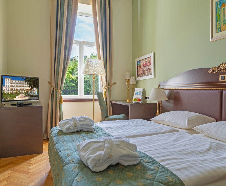Ein helles Hotelzimmer mit zwei Einzelbetten, einem Fernseher und einem Fenster mit Blick ins Freie. Die Wände sind in sanften Grüntönen gestrichen, und es gibt eine gemütliche Atmosphäre mit einem Teppichboden und dekorativen Elementen.