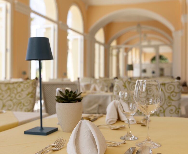 Ein elegantes Restaurant mit hellen Wänden und großen Fenstern. Tische sind mit gelben Tischdecken gedeckt, dekoriert mit gefalteten Servietten, Gläsern und Besteck. Eine kleine Pflanze und eine Tischlampe ergänzen das stilvolle Ambiente.