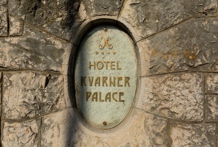 Schild des Hotels Kvarner Palace, eingebettet in eine steinerne Wand. Das Schild hat eine ovale Form und zeigt den Hotelnamen in goldener Schrift. Die Umgebung wirkt rustikal und einladend. Schild des Hotels Kvarner Palace, eingebettet in eine steinerne Wand. Das Schild hat eine ovale Form und zeigt den Hotelnamen in goldener Schrift. Die Umgebung wirkt rustikal und einladend.