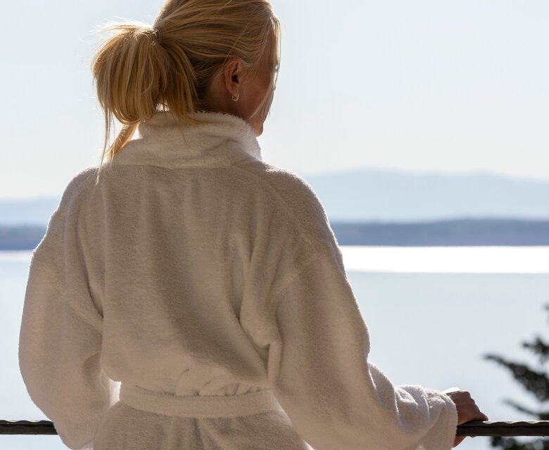 Eine Person in einem weißen Bademantel steht auf einem Balkon und blickt auf einen ruhigen See mit Bergen im Hintergrund. Die Atmosphäre ist entspannt und friedlich, ideal für einen Wellnessurlaub.