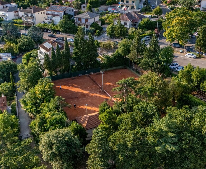 Luftaufnahme einer Wohngegend mit vielen Häusern und Bäumen. Im Vordergrund ist ein Tennisplatz umgeben von Grünflächen zu sehen. Die sanften Hügel im Hintergrund verleihen der Szene eine ruhige Atmosphäre.