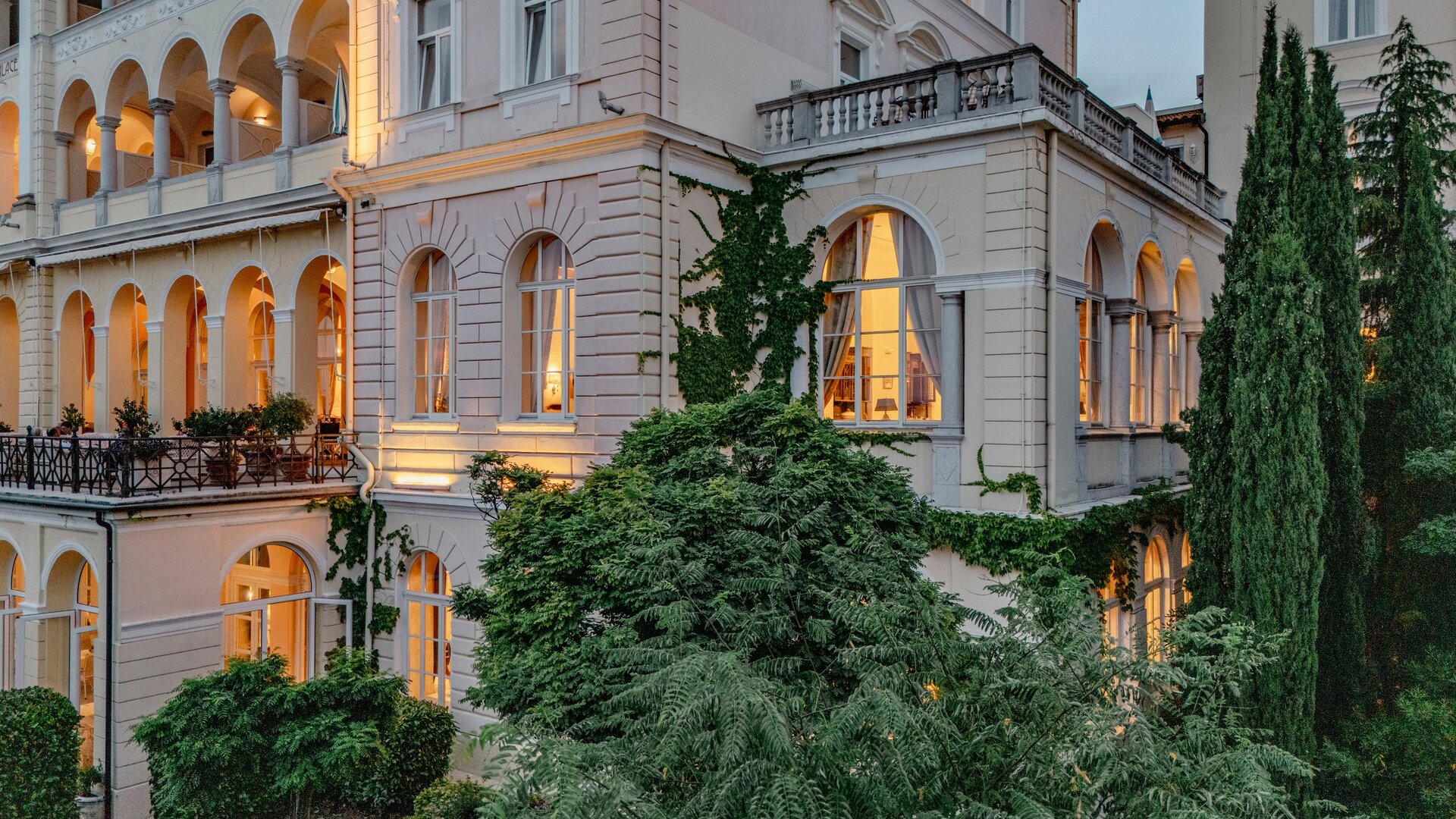 Ein elegantes Hotelgebäude mit großen Fenstern und einem Balkon, umgeben von üppigem Grün. Die warmen Lichter der Fenster schaffen eine einladende Atmosphäre in der Dämmerung.