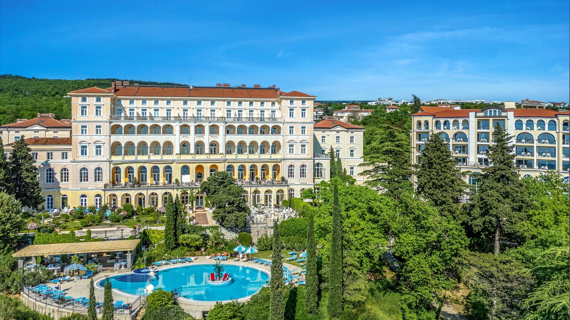 Luxuriöses Hotel mit einem großen Pool inmitten von üppigem Grün. Die Architektur ist elegant, umgeben von Bäumen und Sträuchern, unter einem strahlend blauen Himmel. Ideal für einen entspannten Urlaub.