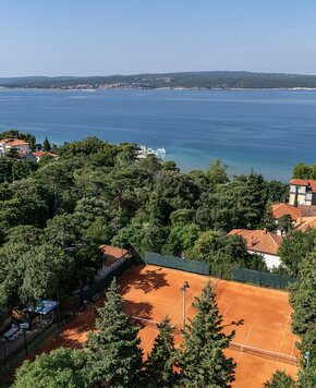 Ein Blick auf einen Tennisplatz mit rotem Sand, umgeben von Bäumen und Villen. Im Hintergrund erstreckt sich ein ruhiger See mit sanften Hügeln und einer kleinen Hafenanlage. Der Himmel ist klar und sonnig.