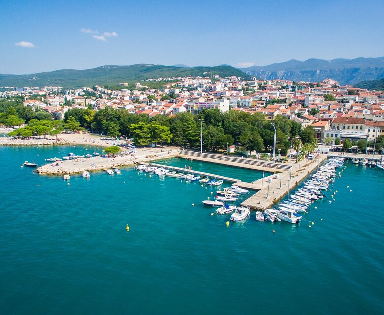 Luftaufnahme eines malerischen Küstenortes mit einem Hafen, umgeben von grünen Bäumen und bunten Gebäuden. Boote liegen im Wasser, während im Hintergrund sanfte Hügel und ein klarer blauer Himmel zu sehen sind.