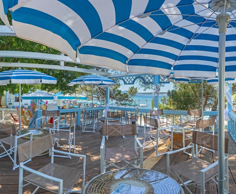 Ein einladender Strandbereich mit blauen und weißen Sonnenschirmen, Holzterrasse und gemütlichen Sitzgelegenheiten. Im Hintergrund ist der Strand und das glitzernde Wasser sichtbar, umgeben von Bäumen und einer entspannten Atmosphäre.