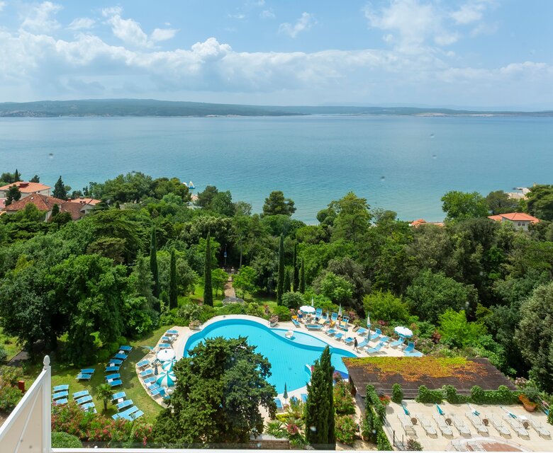 Blick auf einen Pool umgeben von üppigem Grün und Sonnenliegen, mit dem glitzernden Wasser im Hintergrund und einem klaren Himmel. Die Atmosphäre ist entspannt und einladend, ideal für einen Urlaub.