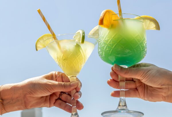 Zwei Hände halten farbenfrohe Cocktails in eleganten Gläsern. Der linke Cocktail ist gelb mit einer Limettenscheibe und einem Strohhalm, der rechte ist grün mit einer Orangenscheibe. Im Hintergrund ist ein klarer Himmel zu sehen.