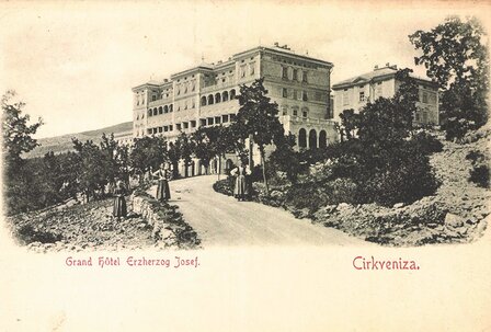 Historisches Grand Hotel Erzherzog Josef, umgeben von Bäumen, mit einer kurvenreichen Straße im Vordergrund. Menschen in zeitgenössischer Kleidung spazieren entlang der Straße. Die Architektur des Hotels ist prächtig und beeindruckend.